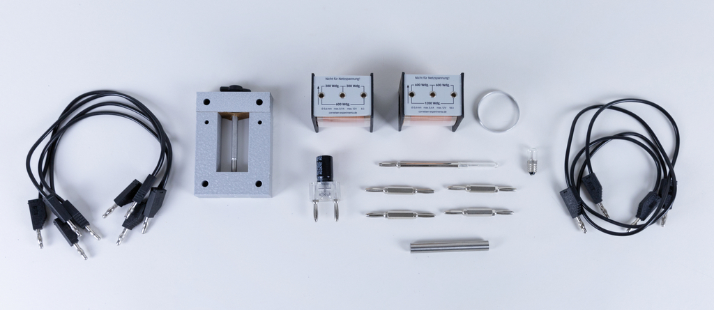 Das Bild zeigt eine Auswahl an Experimentierzubehör, darunter Drähte, zwei Transformatoren, ein Universalträger und verschiedene Metallteile. Diese Komponenten sind für edukative Experimente im Bereich Physik oder Elektrotechnik konzipiert.