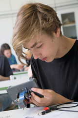 Ein Schüler arbeitet konzentriert an einem technischen Experimentiergerät, in das er verschiedene Komponenten einfügt. Im Hintergrund sind weitere Schüler zu sehen, die ebenfalls mit Experimentiermaterialien beschäftigen.