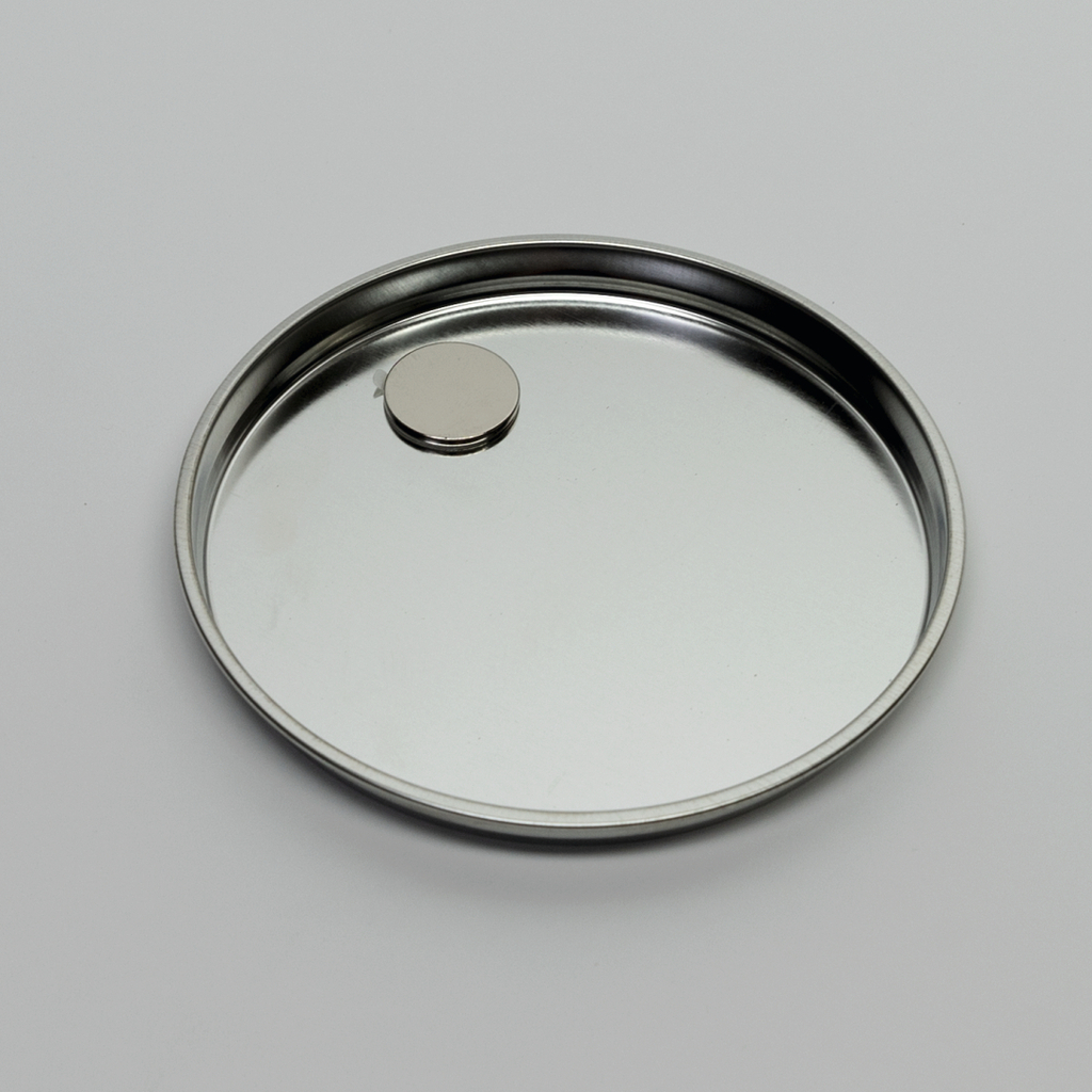 Auf dem Bild ist eine runde, flache Metallschale zu sehen, in deren Mitte sich ein kleiner kreisförmiger Magnet befindet. Die Oberfläche der Schale ist glänzend und reflektiert das Licht.