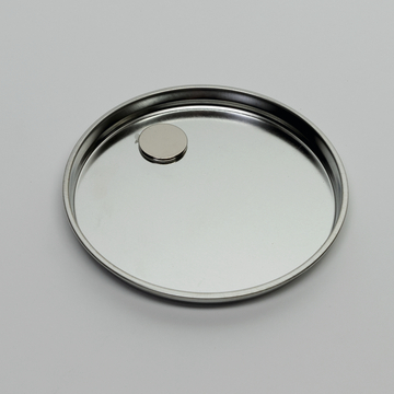 Auf dem Bild ist eine runde, flache Metallschale zu sehen, in deren Mitte sich ein kleiner kreisförmiger Magnet befindet. Die Oberfläche der Schale ist glänzend und reflektiert das Licht.