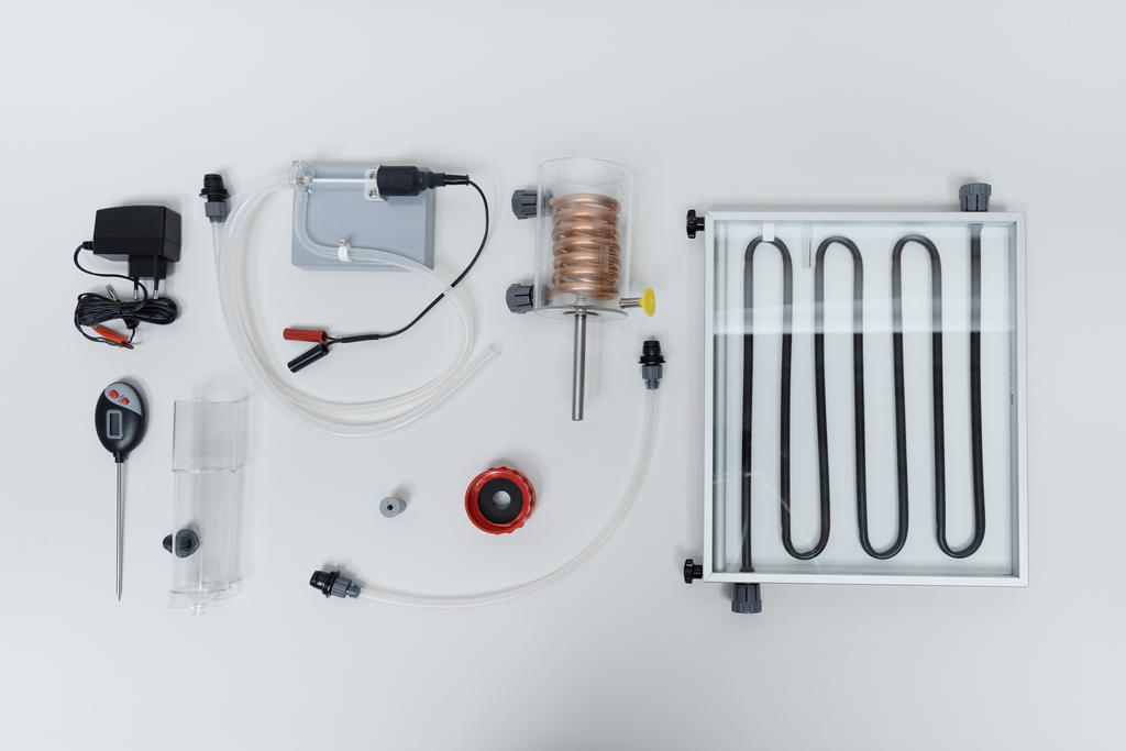 Das Bild zeigt eine Anordnung von Experimentiermaterialien, darunter Rohrtiegel, ein Temperaturfühler, eine Pumpe und Schläuche. Diese Komponenten sind wahrscheinlich für thermodynamische Experimente und Demonstrationen im Bildungsbereich gedacht.
