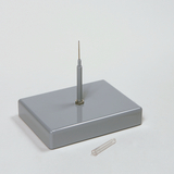 Das Bild zeigt ein graues Experimentiergerät mit einer senkrechten Nadel und einer transparenten Kapsel daneben. Es könnte für wissenschaftliche Experimente im Bildungsbereich genutzt werden, um physikalische oder chemische Konzepte zu demonstrieren.