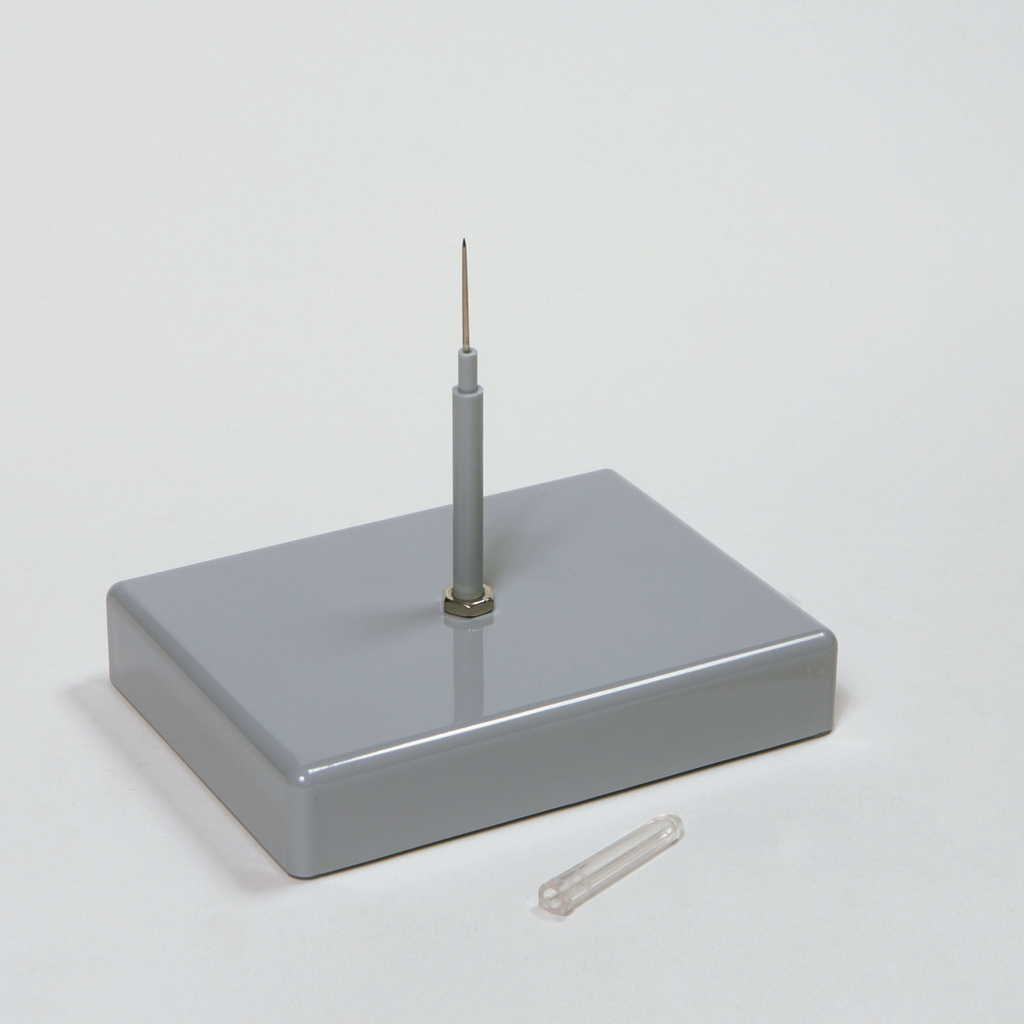 Das Bild zeigt ein graues Experimentiergerät mit einer senkrechten Nadel und einer transparenten Kapsel daneben. Es könnte für wissenschaftliche Experimente im Bildungsbereich genutzt werden, um physikalische oder chemische Konzepte zu demonstrieren.