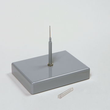 Das Bild zeigt ein graues Experimentiergerät mit einer senkrechten Nadel und einer transparenten Kapsel daneben. Es könnte für wissenschaftliche Experimente im Bildungsbereich genutzt werden, um physikalische oder chemische Konzepte zu demonstrieren.