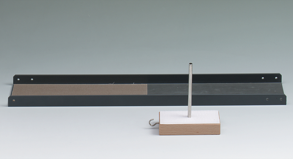Das Bild zeigt eine schwarze Metallbasis mit einer sandigen Fläche und einen vertikalen Stab, der auf einem rechteckigen Holzblock mit einer weißen Oberfläche montiert ist. Dieses Set könnte für physikalische Experimente zur Demonstration von Kräften oder Bewegungen verwendet werden.