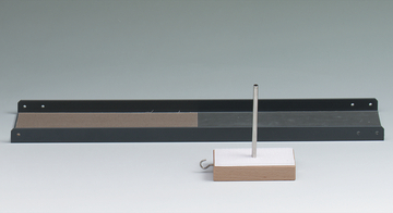 Das Bild zeigt eine schwarze Metallbasis mit einer sandigen Fläche und einen vertikalen Stab, der auf einem rechteckigen Holzblock mit einer weißen Oberfläche montiert ist. Dieses Set könnte für physikalische Experimente zur Demonstration von Kräften oder Bewegungen verwendet werden.