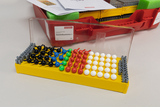 Das Bild zeigt ein Molekülbaukastensystem mit farbigen Kugeln und Stäben auf einer gelben Grundplatte, das zur Veranschaulichung chemischer Strukturen dient. Im Hintergrund ist ein rotes Aufbewahrungsteil mit weiteren Materialien und Anleitungen zu sehen.