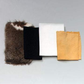 Das Bild zeigt vier verschiedene Textilien: ein Stück Tierfell, ein schwarzes Tuch, ein weißes Baumwollstück und ein orangefarbenes Lederstück. Die Materialien sind möglicherweise für experimentelle Zwecke im Bildungsbereich gedacht.