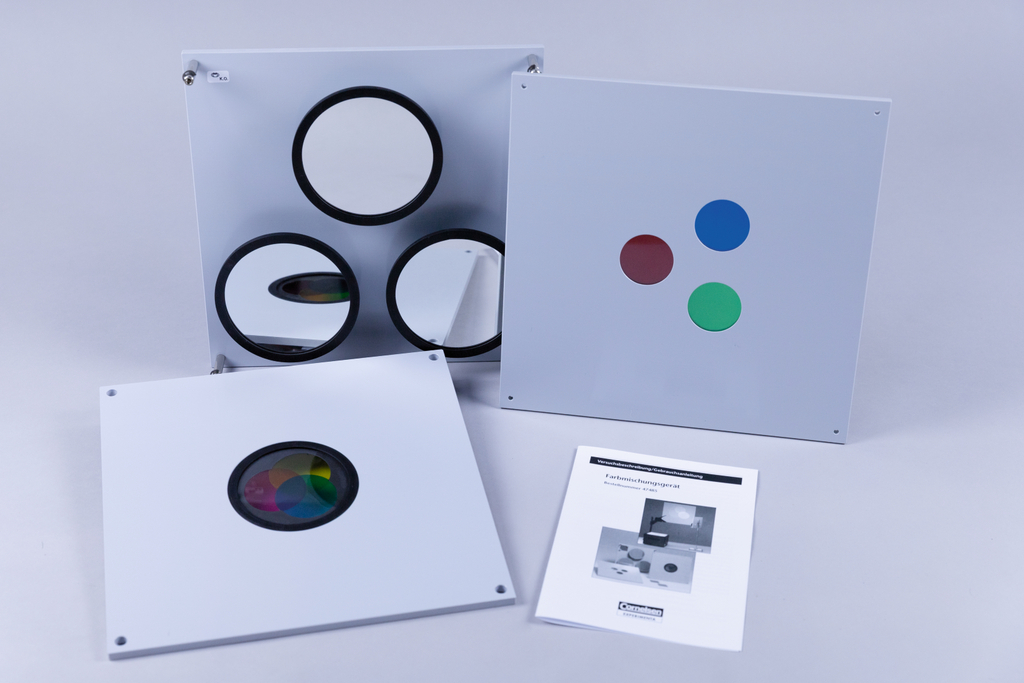 Das Bild zeigt mehrere Experimentierplatten, darunter eine mit farbigen Kreisen (rot, grün, blau) und eine mit drei runden Spiegeln. Außerdem liegt eine Bedienungsanleitung für ein Farbmischergerät neben den Platten.