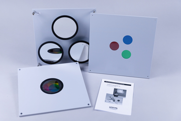 Das Bild zeigt mehrere Experimentierplatten, darunter eine mit farbigen Kreisen (rot, grün, blau) und eine mit drei runden Spiegeln. Außerdem liegt eine Bedienungsanleitung für ein Farbmischergerät neben den Platten.