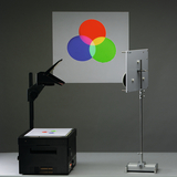 Das Bild zeigt einen Overheadprojektor, der ein Farbdiagramm mit roten, blauen, grünen und gelben Kreisen projiziert. Neben dem Projektor steht eine Halterung, die vermutlich eine transparente Folie oder ein weiteres Experimentiermaterial hält.