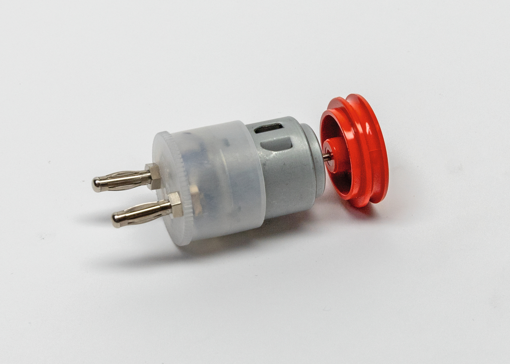 Das Bild zeigt einen elektrischen Motor mit einem transparenten Gehäuse und zwei Anschlussstiften. Neben dem Motor ist ein rotes Antriebsrad abgebildet, das für verschiedene Experimente im Bildungswesen verwendet werden kann.