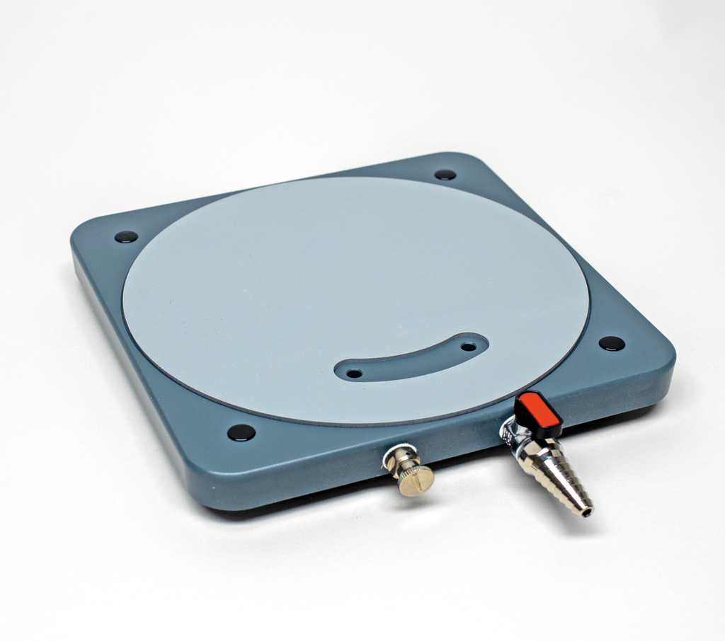 Das Bild zeigt eine graue Experimentierplatte mit einer runden, gehärteten Oberfläche und mehreren Bohrungen. An der Seite befindet sich ein Anschluss mit einem Schalter, vermutlich für eine Luftzufuhr oder andere fluidmechanische Anwendungen.