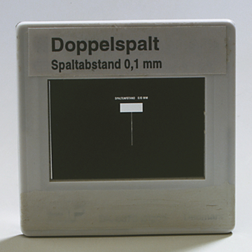 Das Bild zeigt ein Gerät namens "Doppelspalt" mit einem Spaltabstand von 0,1 mm. Es dient dazu, Experimente zur Wellenoptik durchzuführen, indem Licht durch zwei enge Spalte geleitet wird.