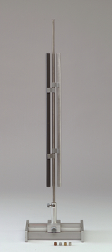 Das Bild zeigt ein Experimentiergerät bestehend aus zwei vertikalen Röhren, einer aus metallischem und einer aus schwarzem Material, die an einem stabilen Basisgestell fixiert sind. Vor der Vorrichtung sind kleine farbige Proben platziert, die wahrscheinlich für Experimente verwendet werden.