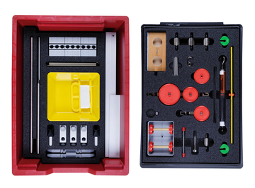 Das Bild zeigt einen Experimentierkasten, der in zwei Fächer unterteilt ist und verschiedene physikalische Komponenten enthält. Die linke Seite beinhaltet unter anderem eine gelbe Schüssel und Stäbe, während die rechte Seite Rollen, Gewichte und weitere Materialien für Experimente bereitstellt.