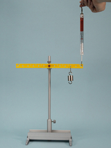 Das Bild zeigt eine experimentelle Vorrichtung zur Durchführung von physikalischen Messungen. Ein Gewicht hängt an einem Hängewaage, die an einer gelben Horizontalstange montiert ist, während ein Handgriff die Waage hält.