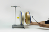 Das Bild zeigt ein physikalisches Experimentiergerät, das aus zwei runden Scheiben und einem Pendel mit einer gelben Kugel besteht. Eine Person schlägt mit einem Stock auf eine der Scheiben, während das Pendel schwingt, um dynamische Kräfte zu demonstrieren.