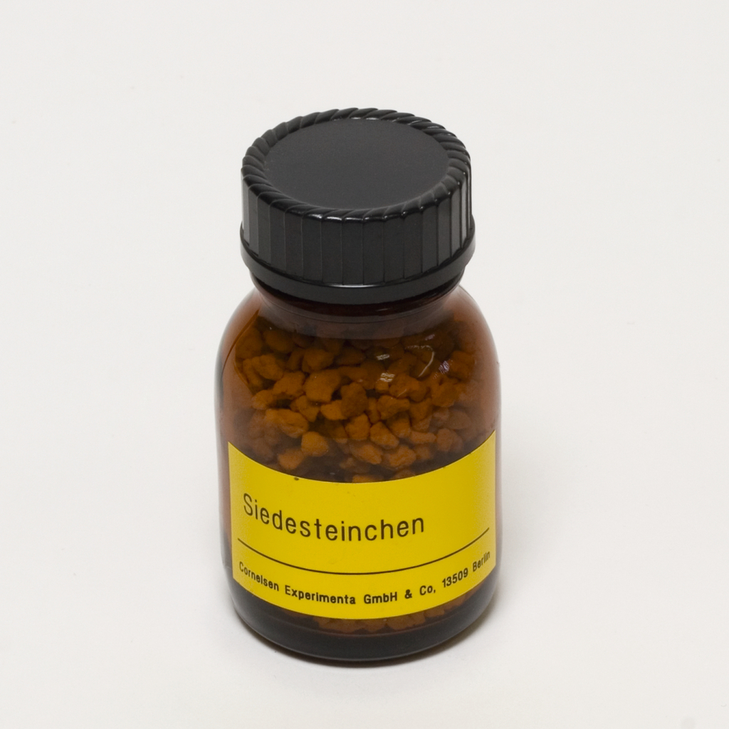 Das Bild zeigt ein braunes Fläschchen mit einer schwarzen Schraubkappe, das mit einem gelben Etikett versehen ist. Das Etikett trägt die Aufschrift „Siedesteinchen“, und die Verpackung enthält kleine, körnige Substanzen, die für chemische Experimente verwendet werden.