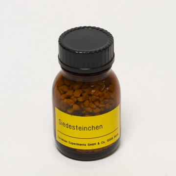 Das Bild zeigt ein braunes Fläschchen mit einer schwarzen Schraubkappe, das mit einem gelben Etikett versehen ist. Das Etikett trägt die Aufschrift „Siedesteinchen“, und die Verpackung enthält kleine, körnige Substanzen, die für chemische Experimente verwendet werden.