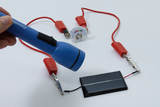 Das Bild zeigt eine experimentelle Anordnung, bei der eine blaue Taschenlampe auf ein Solarpanel gerichtet ist, das mit roten Kabeln verbunden ist. In der Mitte befindet sich ein Messgerät zur Anzeige elektrischer Werte, das die Verbindung und die Funktionsweise des Systems visualisiert.
