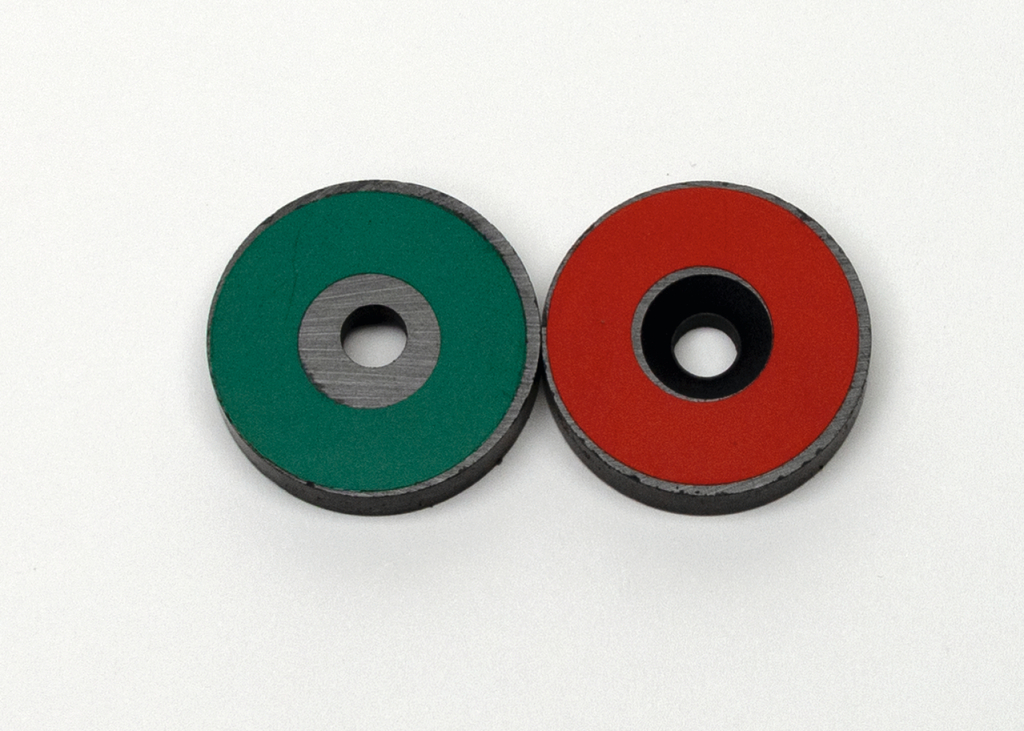 Das Bild zeigt zwei zylindrische Magneten mit einem zentralen Loch, einer ist grün und der andere rot. Diese Magneten sind typischer Experimentierbedarf und eignen sich für verschiedene physikalische Anwendungen im Bildungswesen.