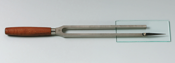 Das Bild zeigt eine Stimmgabel aus Edelstahl mit einem Holzgriff, die auf einer Glasplatte liegt. Dieses Experimentierwerkzeug wird häufig in der Akustik verwendet, um Schwingungen und Tonfrequenzen zu demonstrieren.