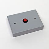 Das Bild zeigt ein graues Gehäuse mit einem roten Druckknopf in der Mitte. Es sind zwei Öffnungen auf der Oberseite sichtbar, die möglicherweise für Anschlüsse oder Sensoren gedacht sind.