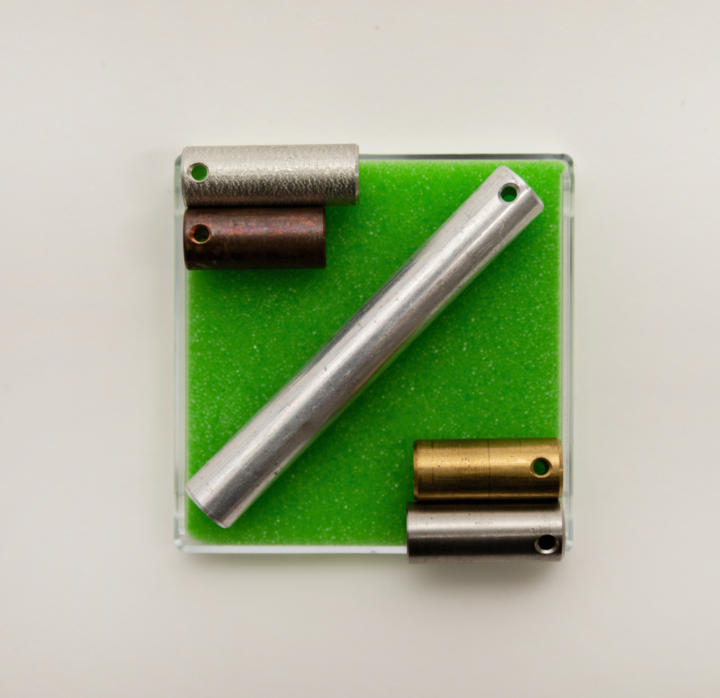 Das Bild zeigt eine Anordnung von verschiedenen zylinderförmigen Objekten aus Metall auf einer grünen Unterlage. Die Objekte haben unterschiedliche Farben und Größen und sind in einer transparenten Box platziert.