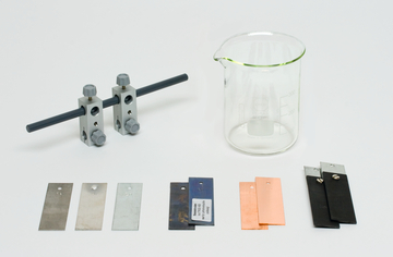 Das Bild zeigt verschiedene experimentelle Materialien, darunter zwei Halterungen mit einer Stange, einen Becher und mehrere Metallplatten in unterschiedlichen Farben und Oberflächen. Diese Materialien sind typischerweise für chemische oder physikalische Experimente im Bildungsbereich vorgesehen.