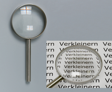 Das Bild zeigt eine Lupe mit einem klaren Glasobjektiv und einem stabilen Metallgriff. Im Hintergrund sieht man wiederholt das Wort "Verkleinern", das durch die Lupe vergrößert wird, was die Funktion des Objekts veranschaulicht.