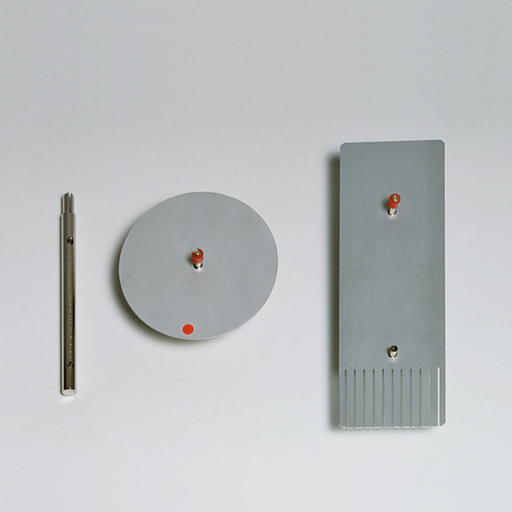 Das Bild zeigt drei experimentelle Geräte aus Metall: einen zylindrischen Stab, eine runde Platte und ein rechteckiges, geripptes Element. Alle Teile sind mit kleinen roten Markierungen versehen und scheinen für physikalische Experimente im Bildungsbereich konzipiert zu sein.