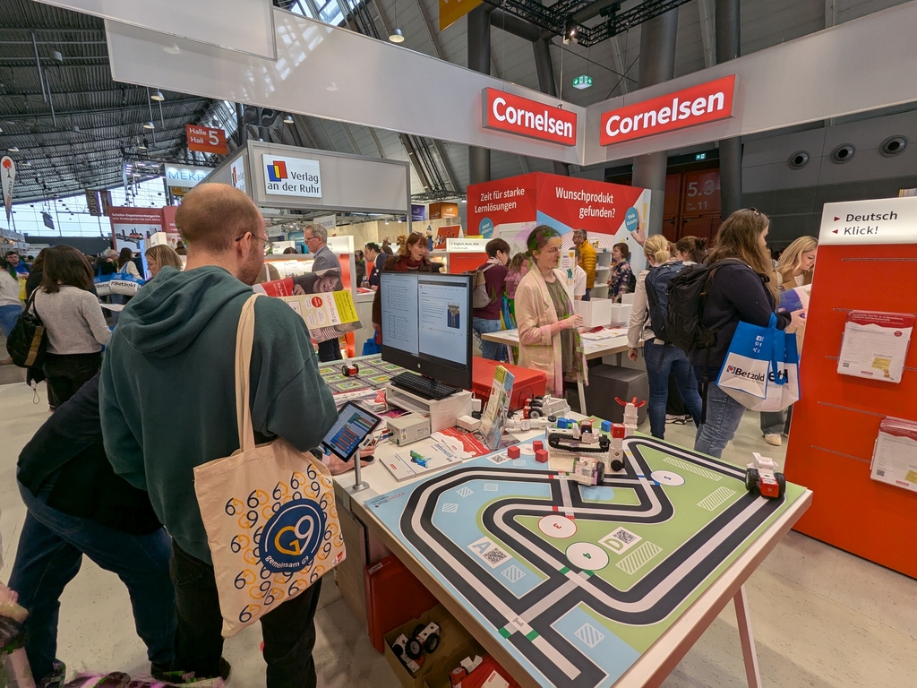 Das Bild zeigt einen Messestand von Cornelsen auf einer Bildungsmesse, wo Besucher an einem interaktiven Experimentierangebot teilnehmen. Auf dem Tisch befindet sich eine Spielmatte mit Fahrzeugen und QR-Codes, während die Besucher Broschüren und Informationen sammeln.