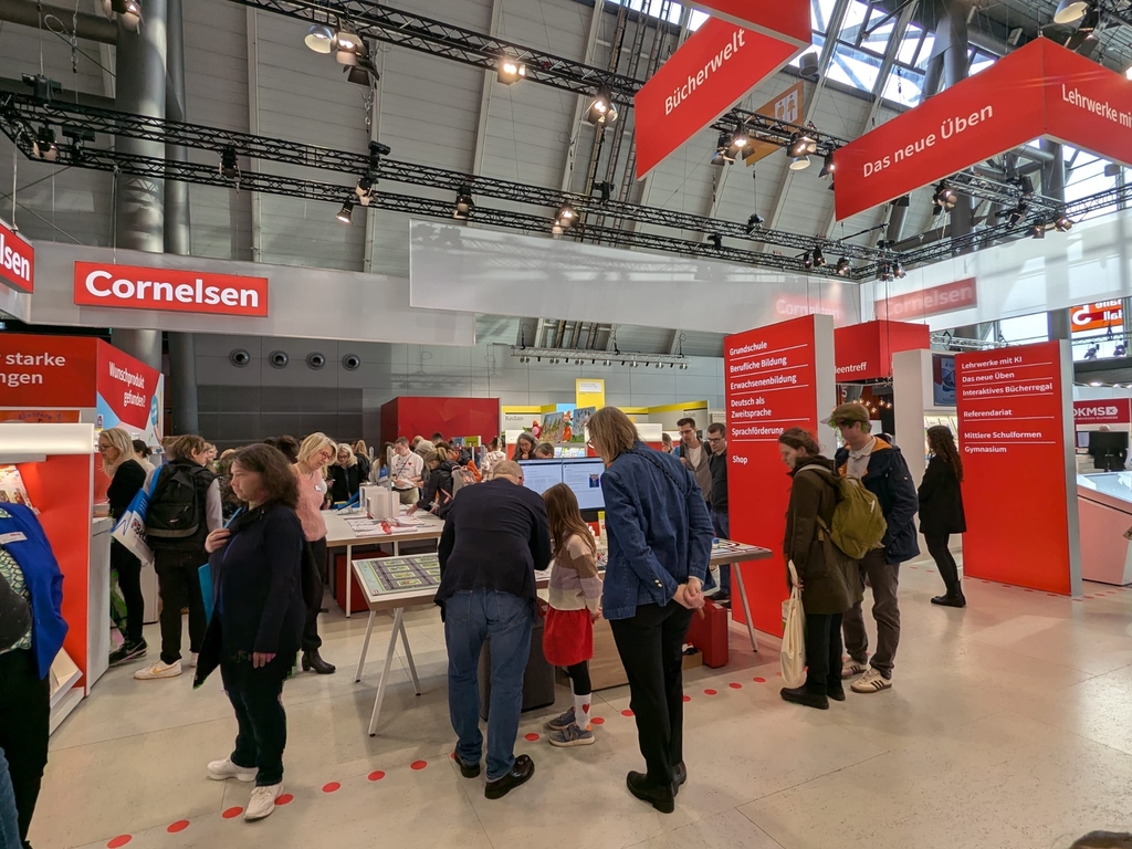 Das Bild zeigt einen belebten Stand von Cornelsen auf einer Bildungsmesse, wo Besucher verschiedene Lernmaterialien und Angebote erkunden. Die Wandtafeln informieren &uuml;ber Produkte f&uuml;r Grundschule, berufliche Bildung und Sprachf&ouml;rderung.