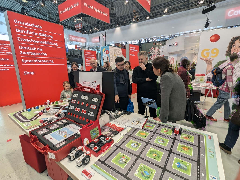 Das Bild zeigt einen Messestand für Experimentierbedarf im Bildungsbereich, mit einem Tisch mit dem eXperiBot Lernroboter für die Grundschule und die Sekundarstufe. Interessierte Besucher, darunter auch Kinder, informieren sich über die Angebote und Dienstleistungen des Standes.