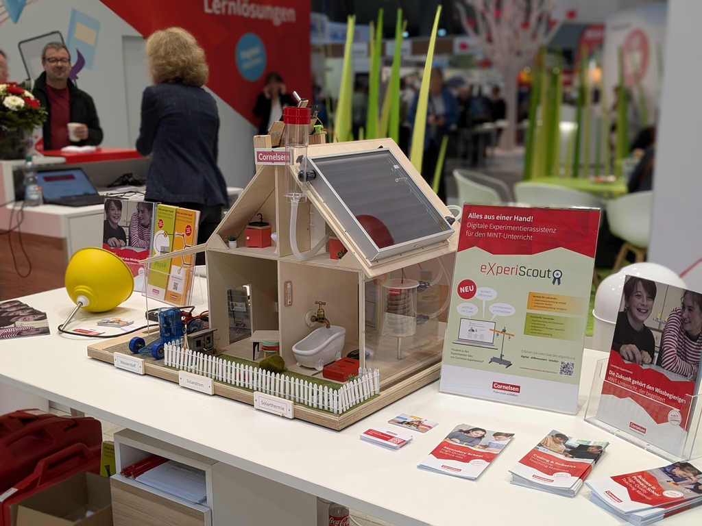 Das Bild zeigt ein Modell eines Energiewandelhauses mit verschiedenen Experimentierm&ouml;glichkeiten, das auf einem Messestand f&uuml;r Bildungsbedarf ausgestellt ist. Daneben stehen Informationsmaterialien zur digitalen Unterst&uuml;tzung im MINT-Unterricht.