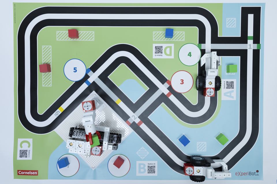 Das Bild zeigt eine Spielmatte mit einer Stra&szlig;enf&uuml;hrung, auf der verschiedene Stationen und farbige Bl&ouml;cke platziert sind. Zwei Roboterfahrzeuge aus Baukl&ouml;tzen sind auf der Matte positioniert und bereit, die Strecke zu erkunden.
