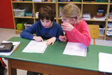 Das Bild zeigt zwei Kinder, die an einem Tisch sitzen und aufmerksam auf Arbeitsblätter schauen. Die Umgebung ist eine Klassenzimmeratmosphäre, mit verschiedenen Materialien und einer Bücherregal im Hintergrund.