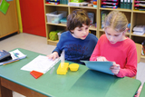 Auf dem Bild sind zwei Kinder an einem Tisch im Klassenzimmer zu sehen. Sie beschäftigen sich mit Experimentiermaterialien, darunter Reagenzgläser und ein Arbeitsblatt, während eines von ihnen ein Tablet betrachtet.