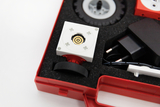Das Bild zeigt ein Experimentier-Kit, das in einem roten Koffer auf Schaumstoffpolstern angeordnet ist. Im Fokus steht ein weißes Modul mit einem zentralen Mechanismus, umgeben von weiteren Komponenten wie Reifen und einem Netzteil.