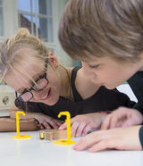 Das Bild zeigt zwei Kinder, die konzentriert auf einen kleinen Holzblock schauen, der zwischen zwei gelben Experimentierständern platziert ist. Sie scheinen begeistert und neugierig zu sein, während sie die experimentellen Materialien untersuchen.