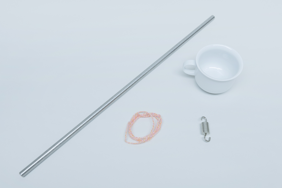 Ein Edelstahlrohr, eine kleine Tasse, ein St&uuml;ck Schnur in rosafarben und ein metallener Wirbel befinden sich auf einer einfarbigen Oberfl&auml;che. Diese Gegenst&auml;nde k&ouml;nnten f&uuml;r physikalische Experimente oder Versuche im Bildungsbereich verwendet werden.