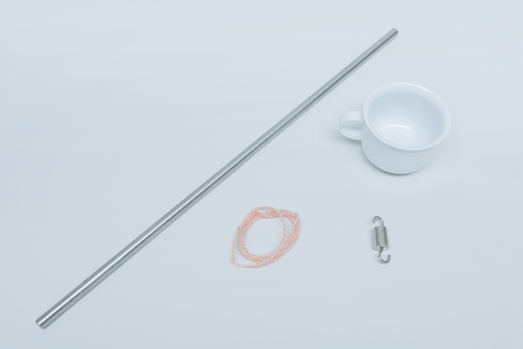 Ein Edelstahlrohr, eine kleine Tasse, ein Stück Schnur in rosafarben und ein metallener Wirbel befinden sich auf einer einfarbigen Oberfläche. Diese Gegenstände könnten für physikalische Experimente oder Versuche im Bildungsbereich verwendet werden.