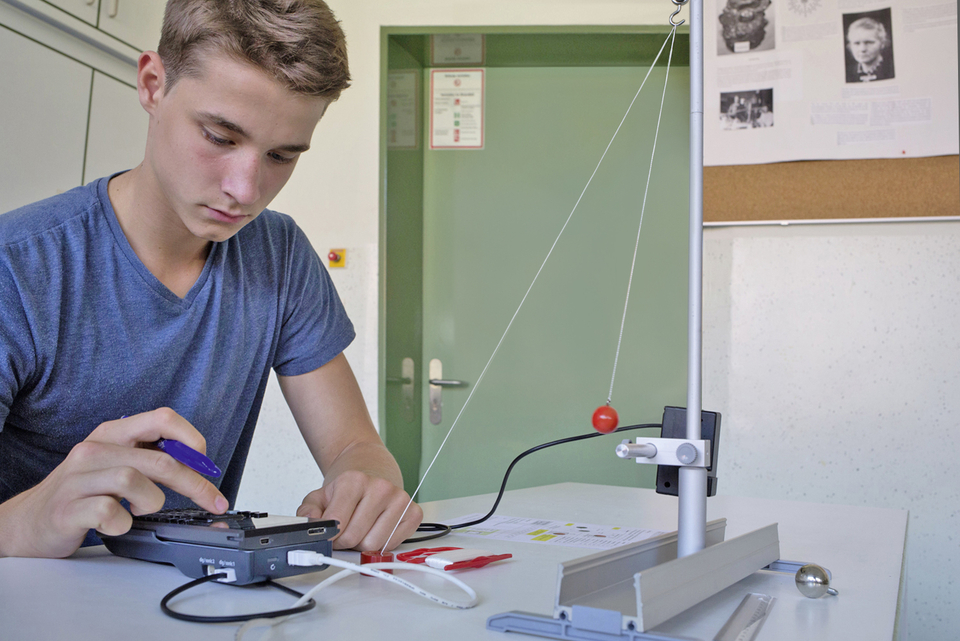 Ein Sch&uuml;ler arbeitet an einem Experiment, das mit einem physikalischen Versuchsaufbau zur Untersuchung von Kr&auml;ften und Bewegungen zu tun hat. Neben ihm liegt ein Ger&auml;t, vermutlich zur Datenerfassung, w&auml;hrend eine rote Kugel an einem Seil schwingt.