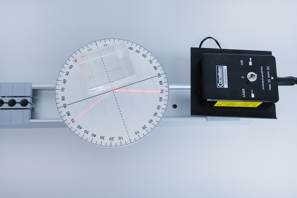 Auf der Arbeitsfl&auml;che ist ein Laserger&auml;t abgebildet, das einen Lichtstrahl auf eine transparente Platte projiziert. Daneben befindet sich ein halbrunder Messkreis mit graduierter Skala zur Analyse des Lichtverlaufs.