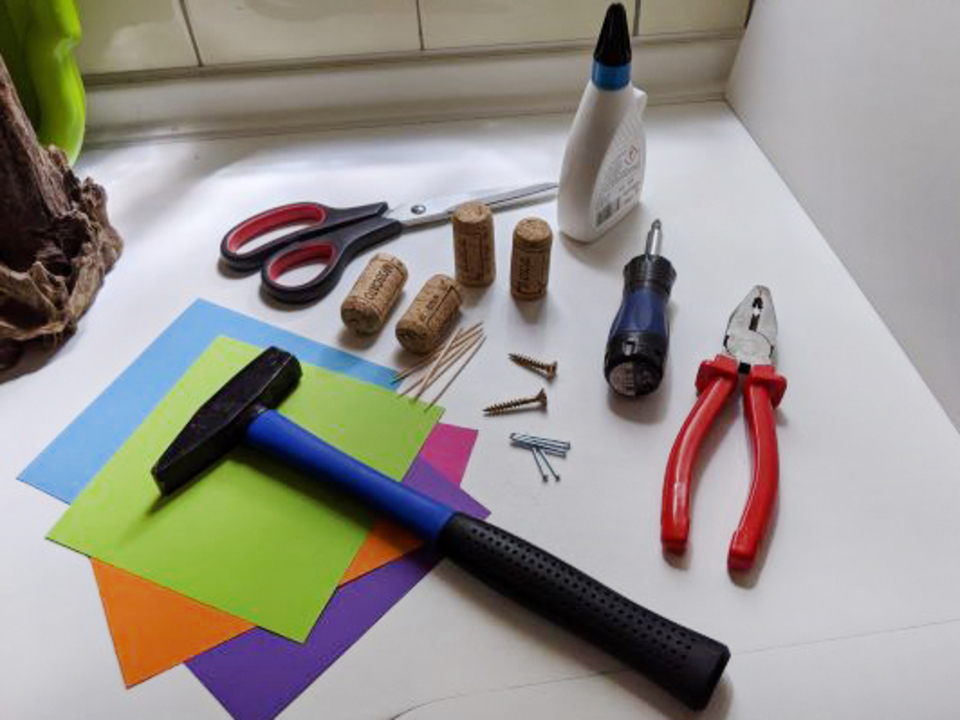 Vielf&auml;ltige Materialien zum Experimentieren sind angeordnet, darunter bunte Bastelpapiere, eine Gummihammer, Scheren, Korken, Schrauben und verschiedene Werkzeuge. Diese Utensilien sind ideal f&uuml;r kreative Projekte im Bildungsbereich.