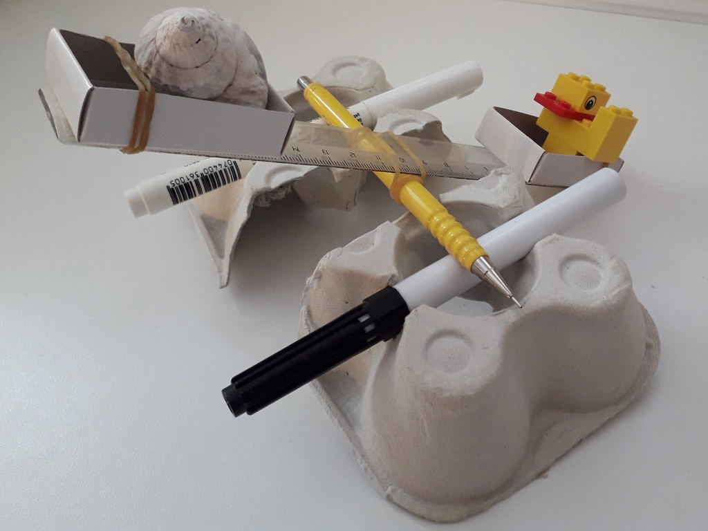 Auf der Unterlage liegen verschiedene Alltagsgegenstände, darunter ein Schneckenhaus, Stifte, ein Gelb-Element aus Bausteinen und ein Lineal, die kreativ angeordnet sind. Die Präsentation vermittelt einen experimentellen und spielerischen Ansatz zur Nutzung von Materialien für Bildungszwecke.