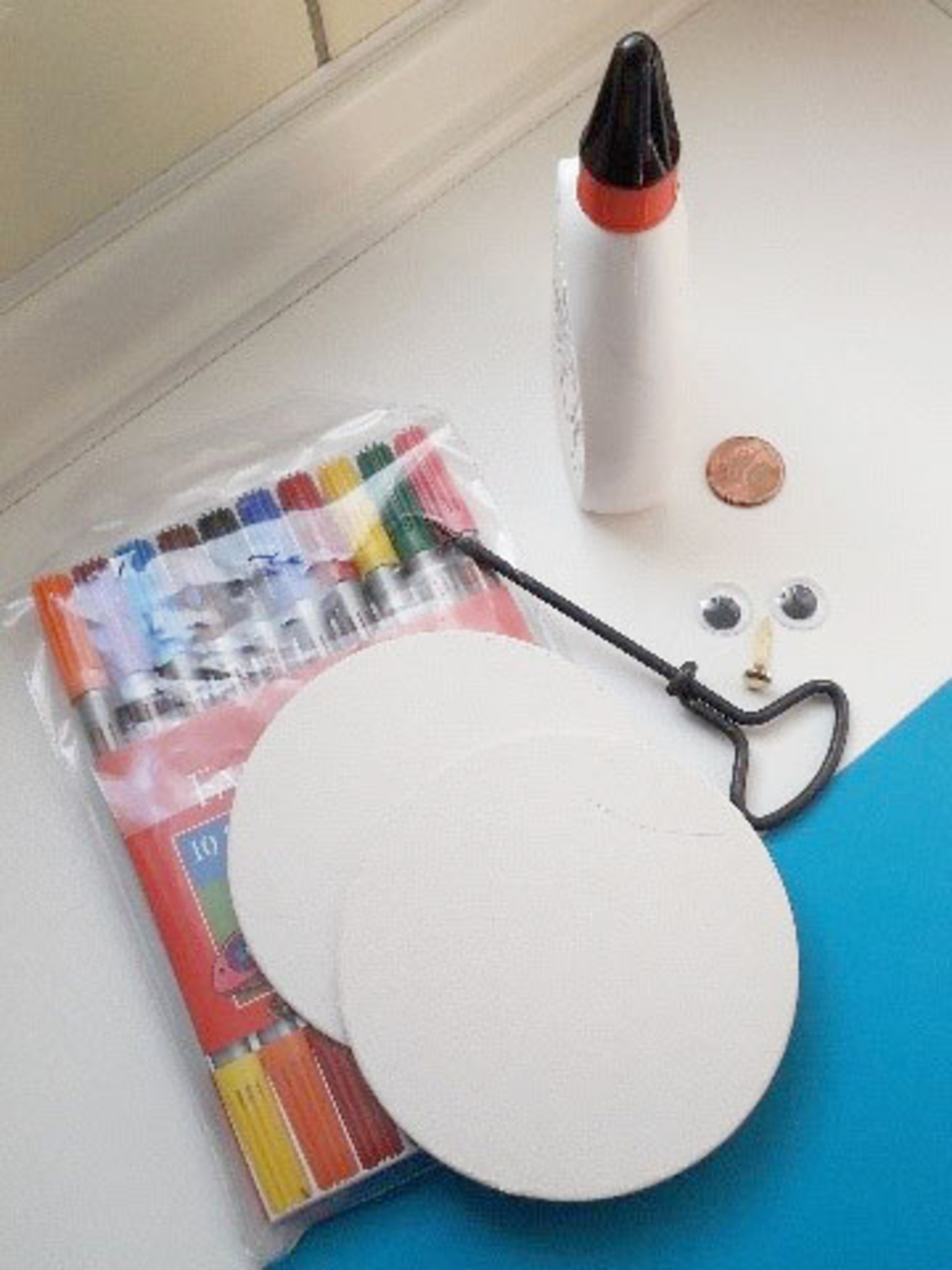 Im Vordergrund sind zwei runde, wei&szlig;e Pappen mit einer Schlaufe, die einen Verbindungspunkt bietet. Daneben befinden sich eine Packung bunter Marker, eine Flasche Kleber, ein centst&uuml;ck und zwei Augen, die f&uuml;r kreatives Basteln verwendet werden k&ouml;nnen.