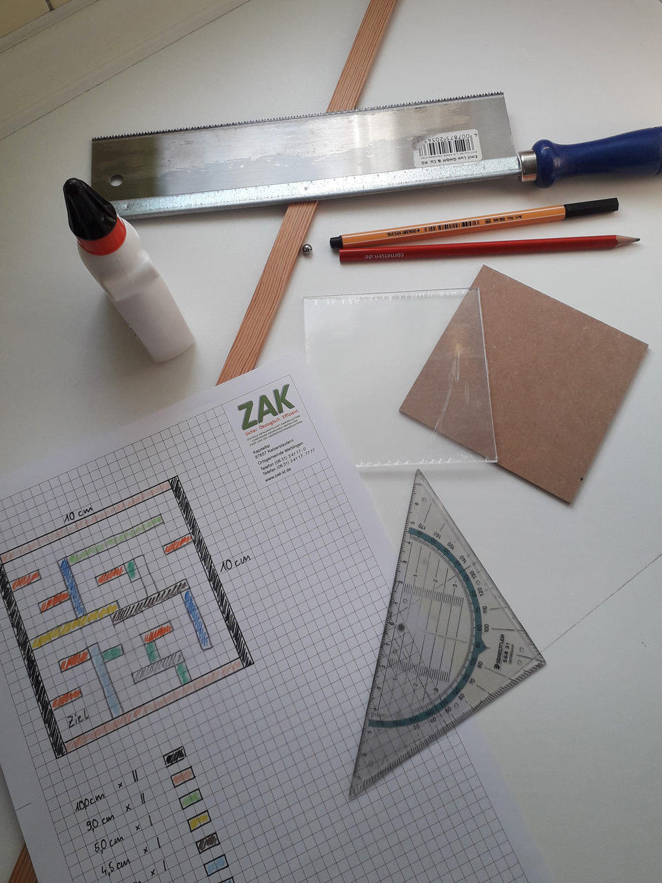 Auf dem Tisch liegen verschiedene Werkzeuge und Materialien, darunter eine Hands&auml;ge, ein Lineal, Kleber, Bleistifte, ein Geodreieck und Zeichenmaterialien. Zudem befindet sich eine Planungsskizze auf kariertem Papier, die eine Bauanleitung zeigt.