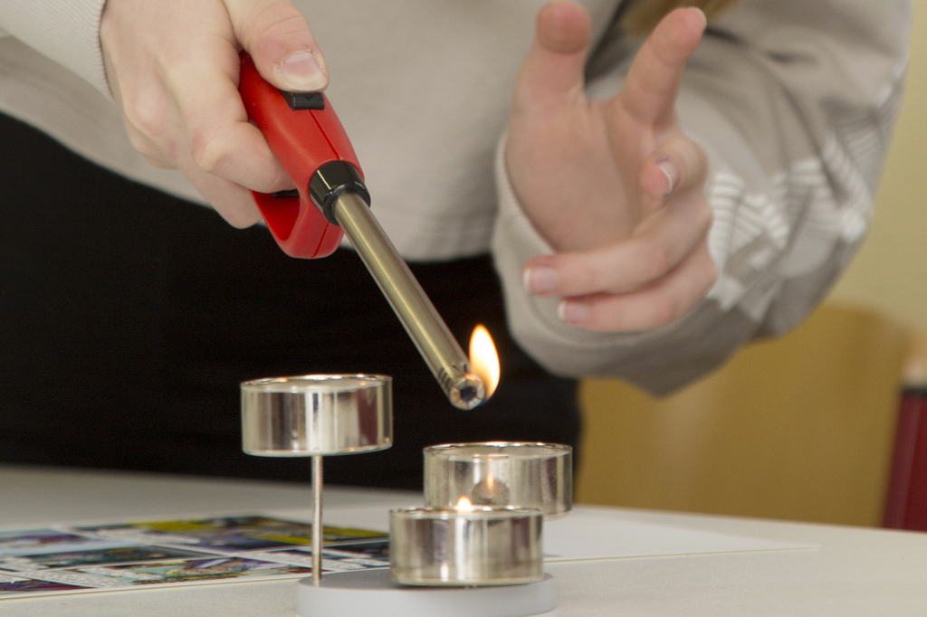 Eine Person verwendet ein Feuerzeug, um eine Kerze anzuzünden, während sie dabei eine Handgeste macht. Auf dem Tisch liegen weitere Teelichter und Materialien für Experimente, die für den Bildungsbereich geeignet sind.